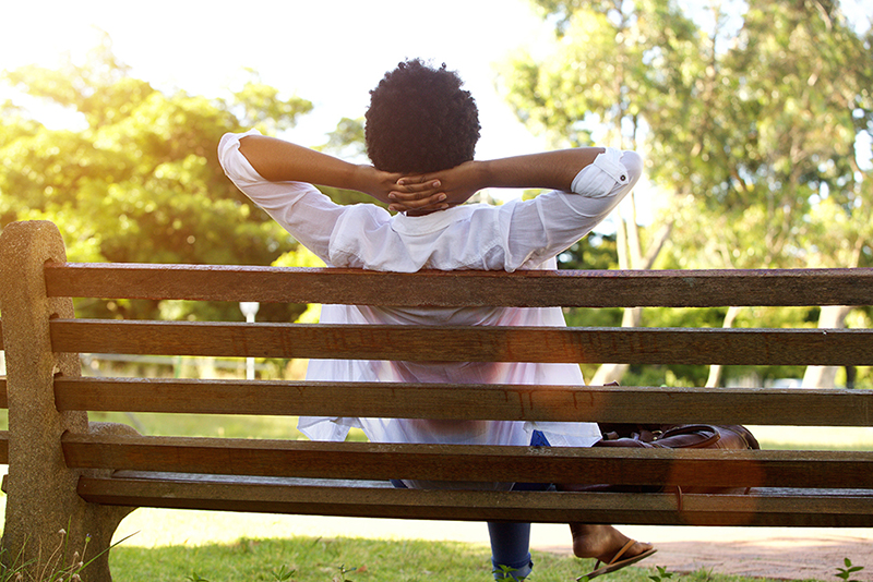 Rear portrait of young lady sitting relaxed on a park bench with hands behind head